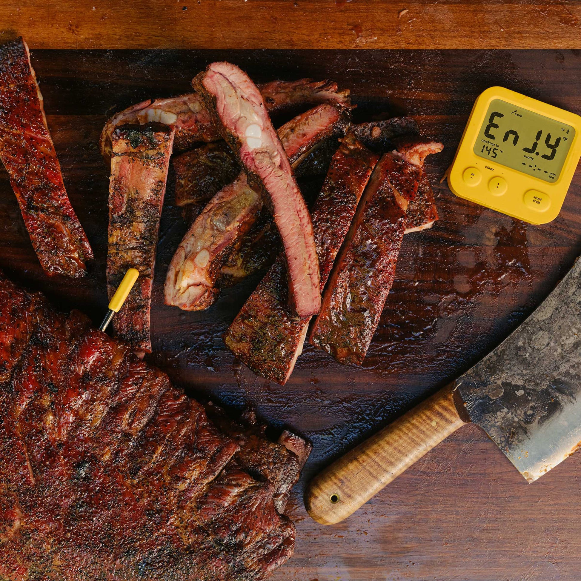 Delicious ribs sliced up on a cutting board, shown with a cleaver and the Combustion Display showing Enjoy and the predictive thermometer inserted into a whole rack of ribs