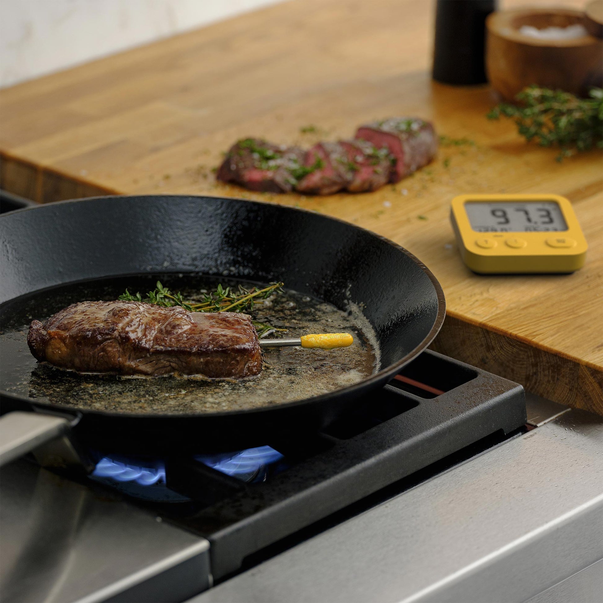 Cooking steak in a pan with the Combustion Predictive Thermometer and Display, using a carbon steel pan on a gas range. Looks delicious. You can see a sliced, perfectly-cooked steak in the background. 
